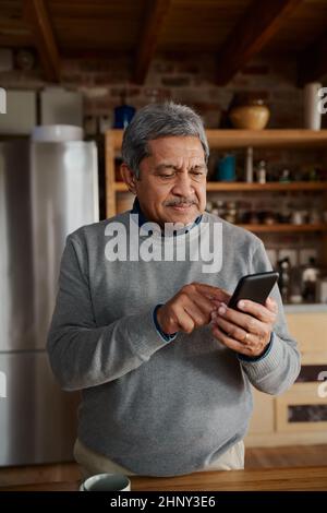 Portrait-Orientierung von glücklichen multikulturellen älteren Männern, die eine Nachricht auf dem Smartphone tippen, während sie in der modernen Küche stehen. Stockfoto