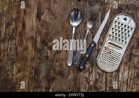 Alte rostige Küchenreibe, Messer, Löffel, Gabel gegen das verfaulende Brett. Küchenutensilien aus Metall. Draufsicht. Selektiver Fokus. Stockfoto