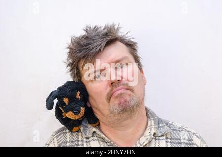 Porträt eines humorvollen Mannes mit einem Hundespielzeug auf der Schulter. Ein Rüde mit zerzauste, verworrene Haare drückt seine Wange gegen das Stofftier. In Innenräumen. Sele Stockfoto