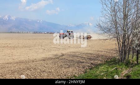 Tractor Pulling heavy metal Walze über trockenes Gebiet mit Bergen, wenig Schnee oben, im Hintergrund. Frühling Feld Vorbereitung. Stockfoto