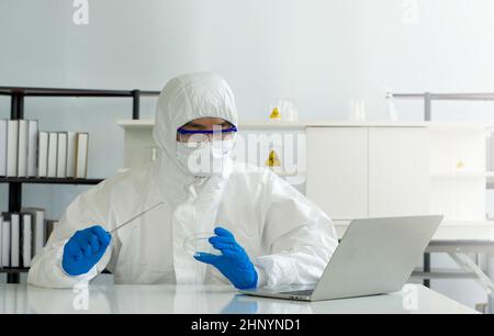 Epidemiologische Forscher in Virusschutzkleidung betrachten den Bildschirm des Laptops, während sie die Nadel und die chemische Petrischale halten. COV Stockfoto