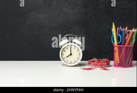 Weißer runder Wecker und ein Metallglas mit Stiften, Bleistiften und Filzstiften auf dem Hintergrund einer leeren schwarzen Kreidetafel, Kopierfläche Stockfoto