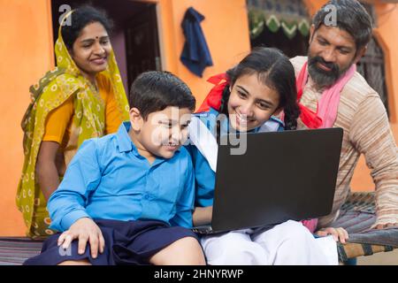 Glückliche junge indische Jungen und Mädchen mit Laptop. Kinder tragen Schuluniform halten Computer mit ihren Eltern hinter ihnen, Online-Bildung und zu Hause s Stockfoto