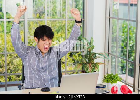 Ja! Glücklich aufgeregt asiatischen Mann hob seinen Arm bis zu feiern Erfolg. Junger Geschäftsmann mit Laptop-Computer an der Büroschreibtisch er froh, wieder Stockfoto