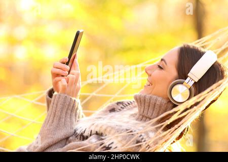 Fröhliche Frau, die im Herbst Kopfhörer trägt und in der Hängematte auf dem Smartphone nachhört Stockfoto