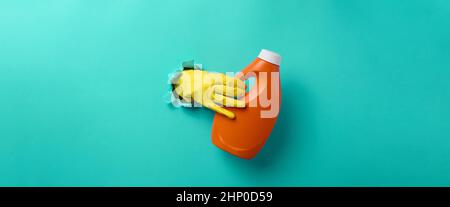 Eine Hand in einem gelben Latex-Reinigungshandschuh hält eine orangefarbene Plastikflasche mit Flüssigwaschmittel vor einem hellblauen Hintergrund. Banner Stockfoto