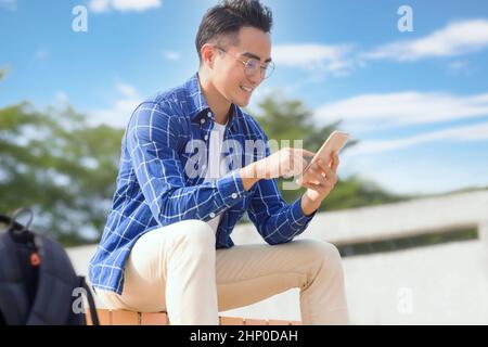 Lächelnder junger asiatischer Mann, der auf Treppen sitzt und Smartphone benutzt Stockfoto