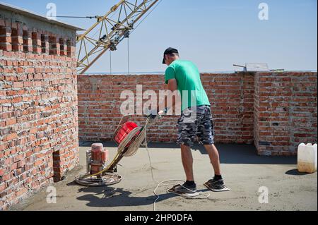 Die ganze Länge des Baumeisters, der die Kelle-Maschine benutzt, während er den Fußboden im neuen Gebäude unter blauem Himmel stachelt. Männlicher Arbeiter, der auf der Baustelle eine Betonoberfläche mit einer Bodentrichschleifmaschine fertig stellte. Stockfoto