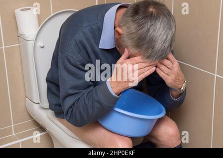 Sowohl Durchfall als auch Erbrechen, Übelkeit, Mann auf der Toilette sitzend, Toilette, eine Schüssel haltend, sich krank fühlend, Kopf in den Händen, krank, unwohl Stockfoto