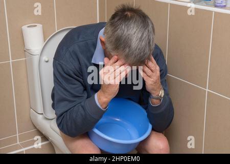 Sowohl Durchfall als auch Erbrechen, Übelkeit, Mann auf der Toilette sitzend, Toilette, eine Schüssel haltend, sich krank fühlend, Kopf in den Händen, krank, unwohl Stockfoto