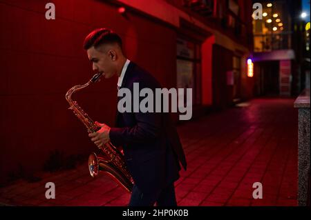 Der junge Saxophonist spielt nachts auf der Straße, urbane Talente. Musiker mit Saxophon im Freien, musikalische Darbietung in der Stadt, Jazzmusiker Stockfoto