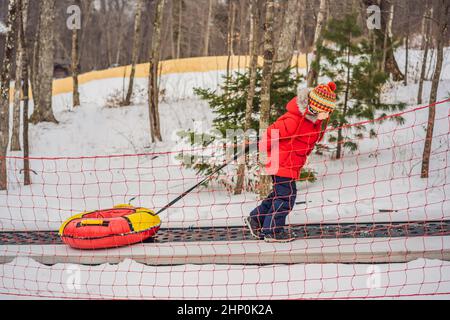 Junge mit Schlauch steigt auf einem Travelator auf den Berg. Kind hat Spaß an der Schneeröhre. Junge reitet einen Schlauch. Winterspaß für Kinder Stockfoto