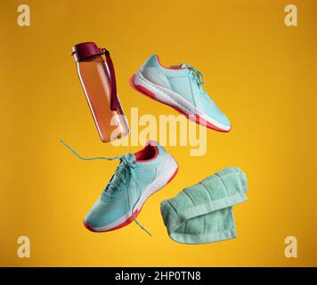Pinke Wasserflasche aus Kunststoff, grünes Handtuch und ein Paar blaue Turnschuhe mit Schnürsenkeln schweben auf gelbem Hintergrund. Schuhe für Sport, Joggen Stockfoto
