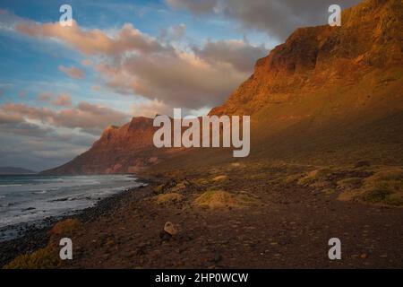 Sonnenuntergang an den Klippen in der Nähe von Famara Beach, Lanzarote, Spanien. Einige Wolken in einem blauen Himmel. Das letzte Sonnenlicht scheint auf den Gipfeln der Berge. Stockfoto