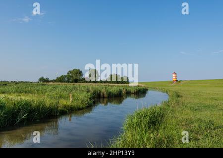 Grüne, idyllische Deichlandschaft vor einem klaren blauen Himmel am Leuchtturm Pilsum mit einem Flutbach im Vordergrund, Pilsum, Krummhoern, Ostfreitag Stockfoto