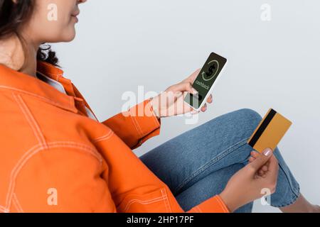 Beschnittene Ansicht einer Frau, die ein Smartphone mit Cashback auf dem Bildschirm verwendet und die Kreditkarte auf grau isoliert hält Stockfoto