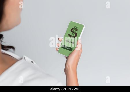 Beschnittene Ansicht einer verschwommenen Frau, die ein Mobiltelefon mit Cashback- und Dollarzeichen auf dem Bildschirm hält, isoliert auf Grau Stockfoto