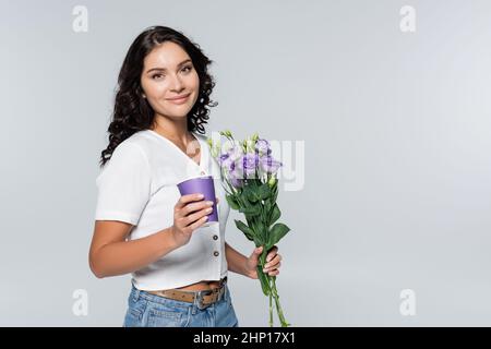 Zufrieden junge Frau hält Bouquet von lila Blumen und Papierbecher isoliert auf grau Stockfoto