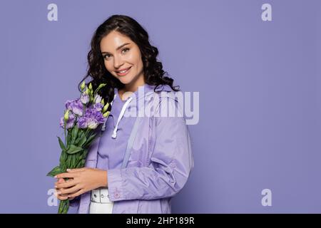 Glückliche junge Frau im Regenmantel mit Blumenstrauß auf lila isoliert Stockfoto