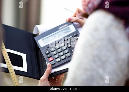 Unkenntliche Frau in Oberbekleidung, die mit einem Taschenrechner Berechnungen anstellt und Notizen auf dem Notizbuch mit Klebeband misst. Stockfoto