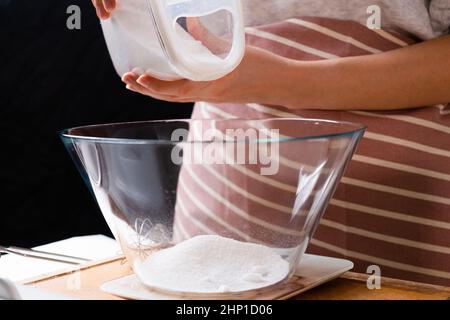 Nahaufnahme Frau Gießen Mehl in eine Rührschüssel für die Vorbereitung von Teig auf dem schwarzen Hintergrund. Selbstgemachter Sauerteig von Grund auf. Stockfoto