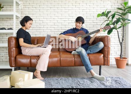 Junge Liebende verbringen im Wohnzimmer Zeit miteinander im Urlaub. Eine asiatische Frau überprüft den Paketzustellungsplan mit einem Laptop, während ihr Freund sie anschaut Stockfoto