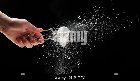 Weibliche Hand hält ein Metallsieb mit Puderzucker. Partikel fliegen in verschiedene Richtungen auf einem schwarzen Hintergrund, sprüchendes Stockfoto