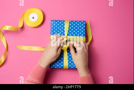 Weibliche Hand bindet ein Seidenband auf einer Geschenkbox, rosa Hintergrund. Vorbereitung auf den Urlaub Stockfoto