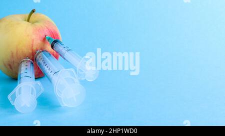 Drei verschiedene medizinische Spritzen, die tief in einen Apfel auf blauem Hintergrund stecken. Die Frucht ist mit Spritzen übersät. Ein reifer roter Apfel, der von al durchbohrt wurde Stockfoto
