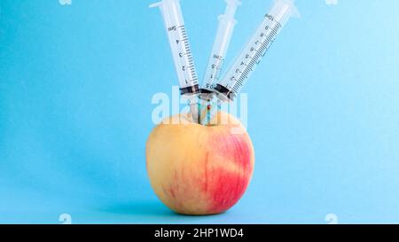 Drei verschiedene medizinische Spritzen, die tief in einen Apfel auf blauem Hintergrund stecken. Die Frucht ist mit Spritzen übersät. Ein reifer roter Apfel, der von al durchbohrt wurde Stockfoto