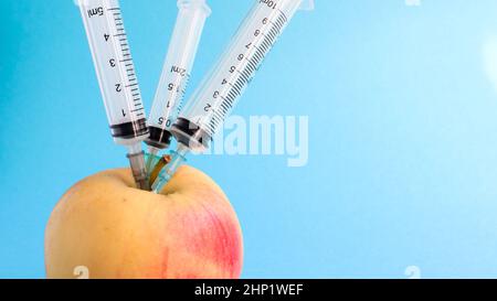 Drei verschiedene medizinische Spritzen, die tief in einen Apfel auf blauem Hintergrund stecken. Die Frucht ist mit Spritzen übersät. Ein reifer roter Apfel, der von al durchbohrt wurde Stockfoto