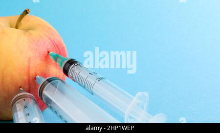 Drei verschiedene medizinische Spritzen, die tief in einen Apfel auf blauem Hintergrund stecken. Die Frucht ist mit Spritzen übersät. Ein reifer roter Apfel, der von al durchbohrt wurde Stockfoto