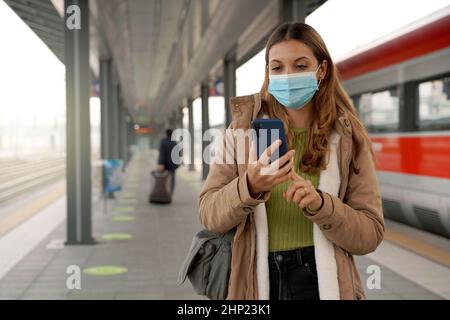 Reisende Frau trägt chirurgische Maske am Bahnhof kaufen Ticket online mit ihrem Handy im Freien Stockfoto