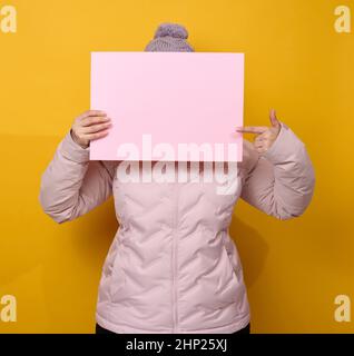 Frau in rosa Winterjacke und Hut hält rosafarbenes Blatt Papier auf gelbem Hintergrund. Saisonaler Verkauf. Platz für die Beschriftung Stockfoto