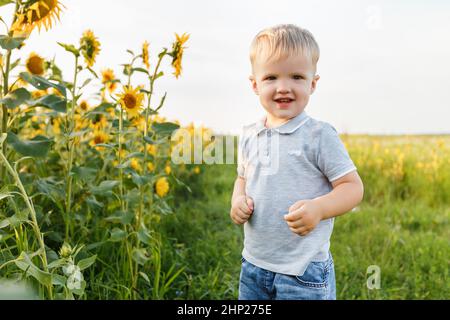 Kleiner Junge, der auf dem Hintergrund der Landschaft spielt und lächelt. Drei Jahre Kind Spaß im Feld der Sonnenblumen am Sonnenuntergang. Sommer im Freien Livestyl Stockfoto