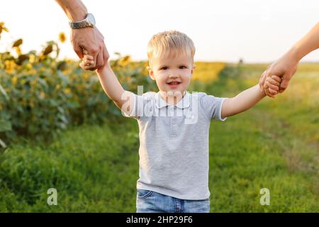 Kleiner Junge lächelt und hält die Hände seiner Eltern auf dem Hintergrund der Landschaft. Drei Jahre Kind mit Familie, die Spaß im Feld der Sonnenblumen auf su Stockfoto