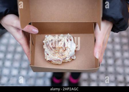 Ein Mädchen hält einen Cinnabon mit Creme in einer Bastelkiste. Lieferung von frischen Backwaren Stockfoto