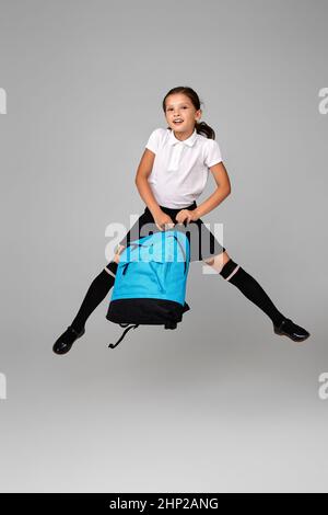 Lächelndes Mädchen mit einem kleinen Schüler, das mit einem blauen Rucksack auf grauem Hintergrund springt. Zurück zur Schule Stockfoto