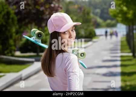 Junge schöne Brünette Skateboarderin Mädchen in Mütze mit Skateboard im Freien im Park stehen und mit Blick auf die Kamera Stockfoto