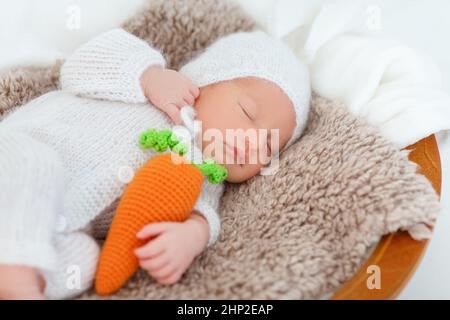 Neugeborenes Baby in weißem Hase-Anzug schlafen auf Holzkorb. Kinderkaninchen mit gestrickter Karotte in den Händen. Kreative Babyfotografie Stockfoto