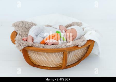 Neugeborenes Baby in weißem Hase-Anzug schlafen auf Holzkorb. Kinderkaninchen mit gestrickter Karotte in den Händen. Kreative Babyfotografie Stockfoto