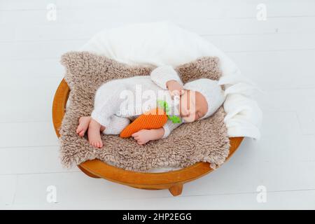 Neugeborenes Baby in weißem Hase-Anzug schlafen auf Holzkorb. Kinderkaninchen mit gestrickter Karotte in den Händen. Kreative Babyfotografie Stockfoto