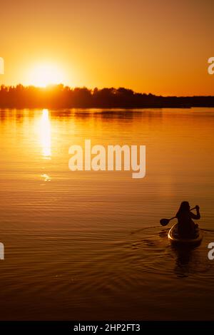 Silhouette einer Frau, die auf einem Paddelbrett sitzt, mit Rudern und Händen, die gerade auf einem wunderschönen See rudern, mit einem erstaunlichen Sonnenuntergang im Hintergrund, der sich darin spiegelt Stockfoto