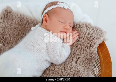 Neugeborenes Baby in weißem Strickanzug schlafen im Holzkorb. Kind ruht sich in niedlicher Pose aus. Kleines Mädchen mit einem zarten Haarreif auf dem Kopf. Kreative Fototrainken für Kleinkinder Stockfoto