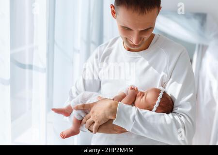 Das neugeborene Baby im weißen Strickanzug schläft auf den Händen des Vaters. Vater und Tochter. Winziges Kind mit einem zarten Haarreif am Kopf. Konzept der glücklichen Elternschaft, lo Stockfoto