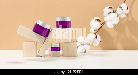 Kosmetikprodukte in einem violetten Glas mit grauem Deckel auf einem hölzernen Podium aus Würfeln, hinter einem Weintraubenzweig mit grünen Blättern. Leer für Marke Stockfoto