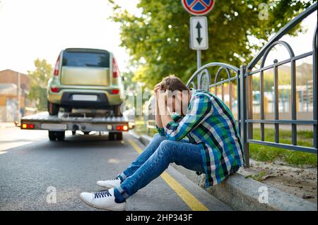 Unglücklicher Mann, der am Straßenrand sitzt und seinen Kopf während der falschen Evakuierung des geparkten Fahrzeugs durch einen Abschleppwagen hält Stockfoto