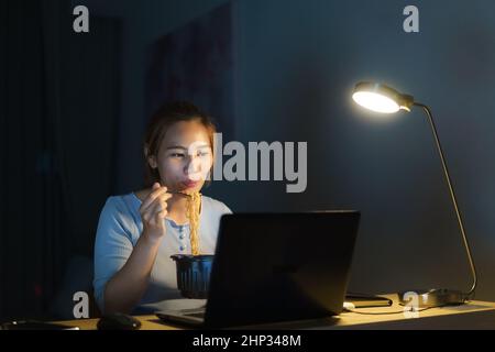 Asiatische Freiberufler intelligente Geschäftsfrau essen Instant Nudeln während der Arbeit auf einem Laptop im Wohnzimmer zu Hause in der Nacht glücklich asiatische Mädchen sitzt auf Stockfoto