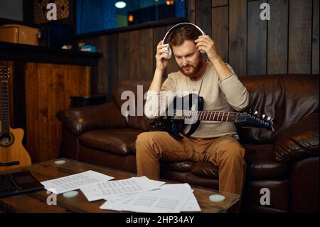 Mann, der Kopfhörer auszieht, beendet den Musikunterricht. Kerl sitzt mit Gitarre zu Hause Studio Stockfoto