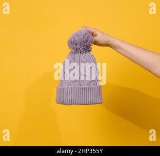 Weibliche Hand hält Strickmütze mit Bommel auf gelbem Hintergrund, warme und bequeme Kleidung Stockfoto